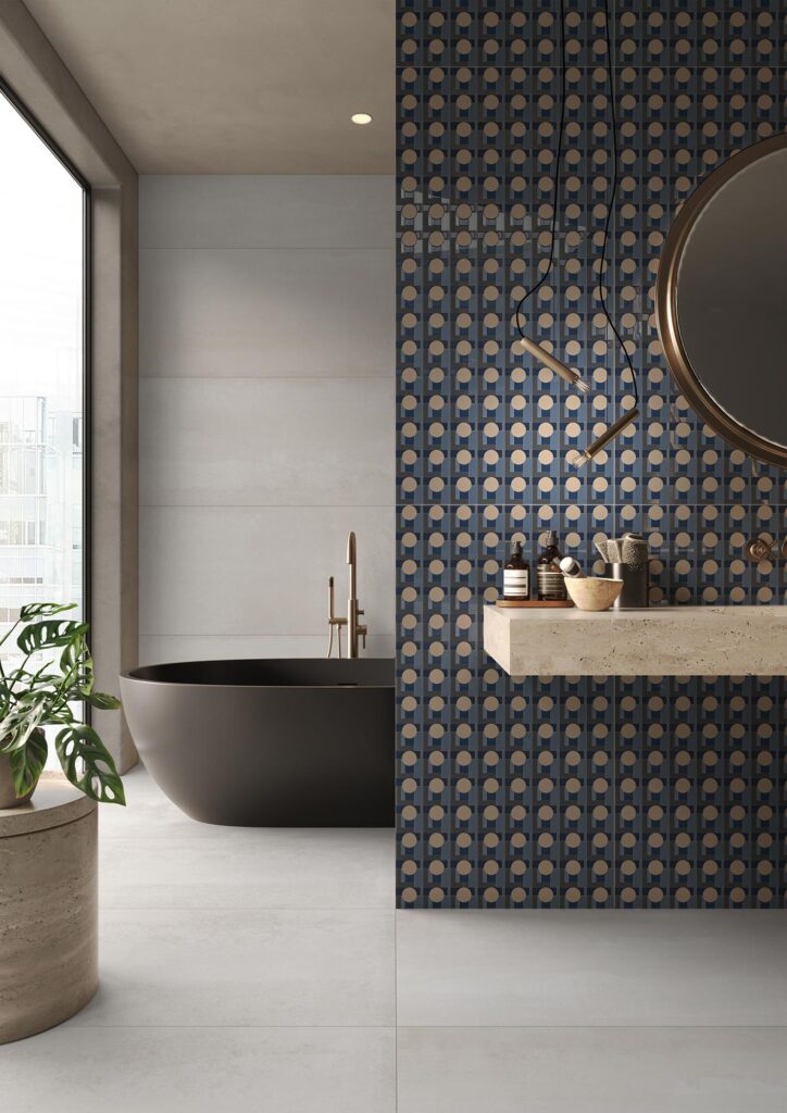 Collezione ART: una raffinata ambientazione bagno di design che mette in mostra la versatilità dei formati 60x120. Sulla parete divisoria a destra è posato il decoro geometrico DotPot 60x120, caratterizzato da un elegante pattern ipnotico nei toni del blu, del grigio e del beige dorato, che fa da sfondo a un lavabo sospeso in pietra e a uno specchio circolare. Il pavimento e la parete di fondo sono rivestiti con le piastrelle Hemisphere Platinum 60x120 dall'effetto cemento chiaro e minimalista. Sullo sfondo, una moderna vasca da bagno freestanding di colore scuro con rubinetteria dorata e una rigogliosa pianta verde completano un'atmosfera lussuosa, luminosa e contemporanea.