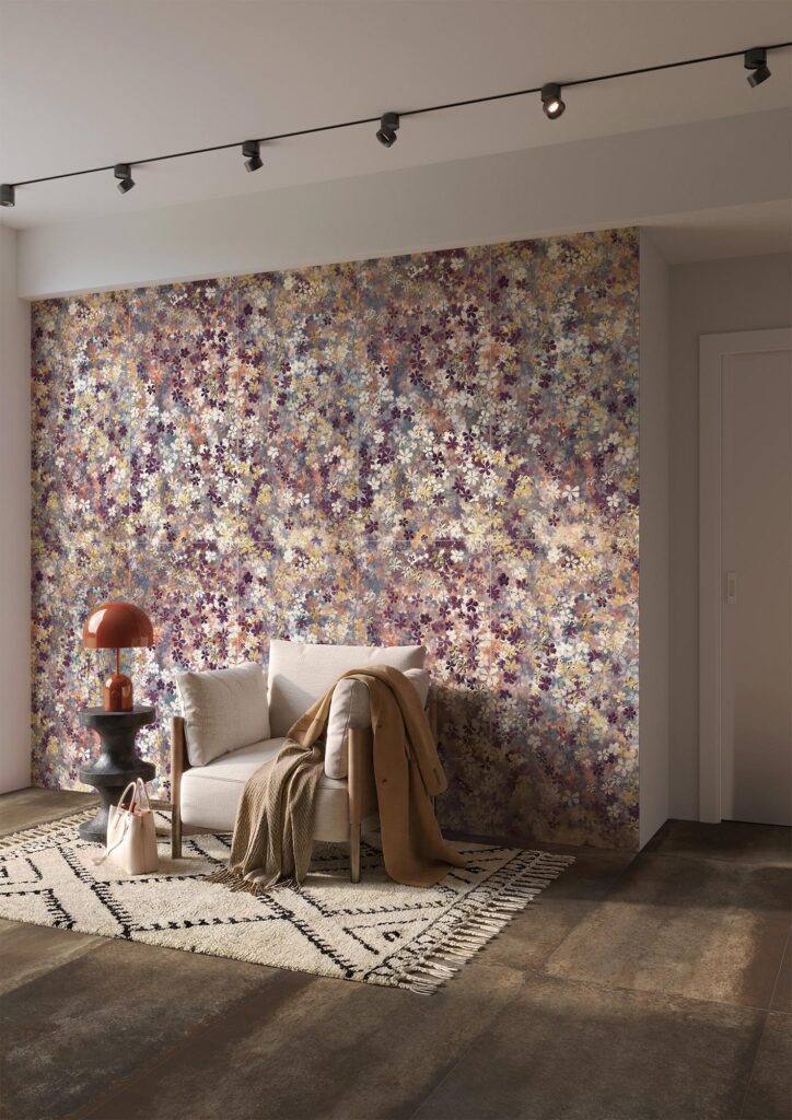 La collezione ART si rivela in questo raffinato ambiente living e ingresso, proponendo a parete i decori floreali Folie nel formato 60x120 cm e a pavimento le superfici metalliche di Hemisphere Copper nel formato 60x120 cm. L'immagine mostra una scenografica parete rivestita con piastrelle ceramiche caratterizzate da un fitto e dettagliato pattern di piccoli fiori nei toni del viola, lilla, bianco e giallo ocra, che creano un ricercato effetto carta da parati. Davanti alla parete, una confortevole poltrona di design in tessuto chiaro, arricchita da una morbida coperta e un cappotto adagiati sopra, è posizionata su un tappeto con motivi geometrici neri e frange. Accanto alla seduta spiccano un tavolino scuro con una lampada da tavolo arancione dal gusto retrò e una borsa poggiata a terra. L'atmosfera dell'ambiente è calda, intima e sofisticata, esaltata dal pavimento in grès scuro effetto metallo e da una moderna illuminazione a faretti orientabili a soffitto.