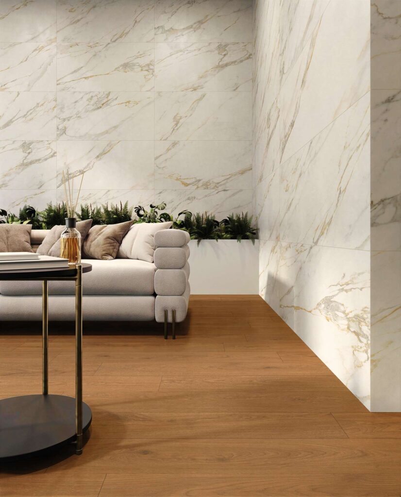 La collezione Elysion definisce un elegante spazio living dove il gres porcellanato effetto legno Barrique Cherry nel formato 20X120 a pavimento incontra il raffinato rivestimento a parete effetto marmo Elysion Calacatta nel formato 60x120. L'ambiente, luminoso e moderno, mostra un angolo salotto con un divano grigio chiaro arricchito da soffici cuscini, una fioriera con piante verdi sullo sfondo e un tavolino di design in primo piano. Le venature calde e dorate della parete effetto marmo si coordinano armoniosamente con le tonalità calde del pavimento in legno, creando un'atmosfera accogliente e di grande prestigio.