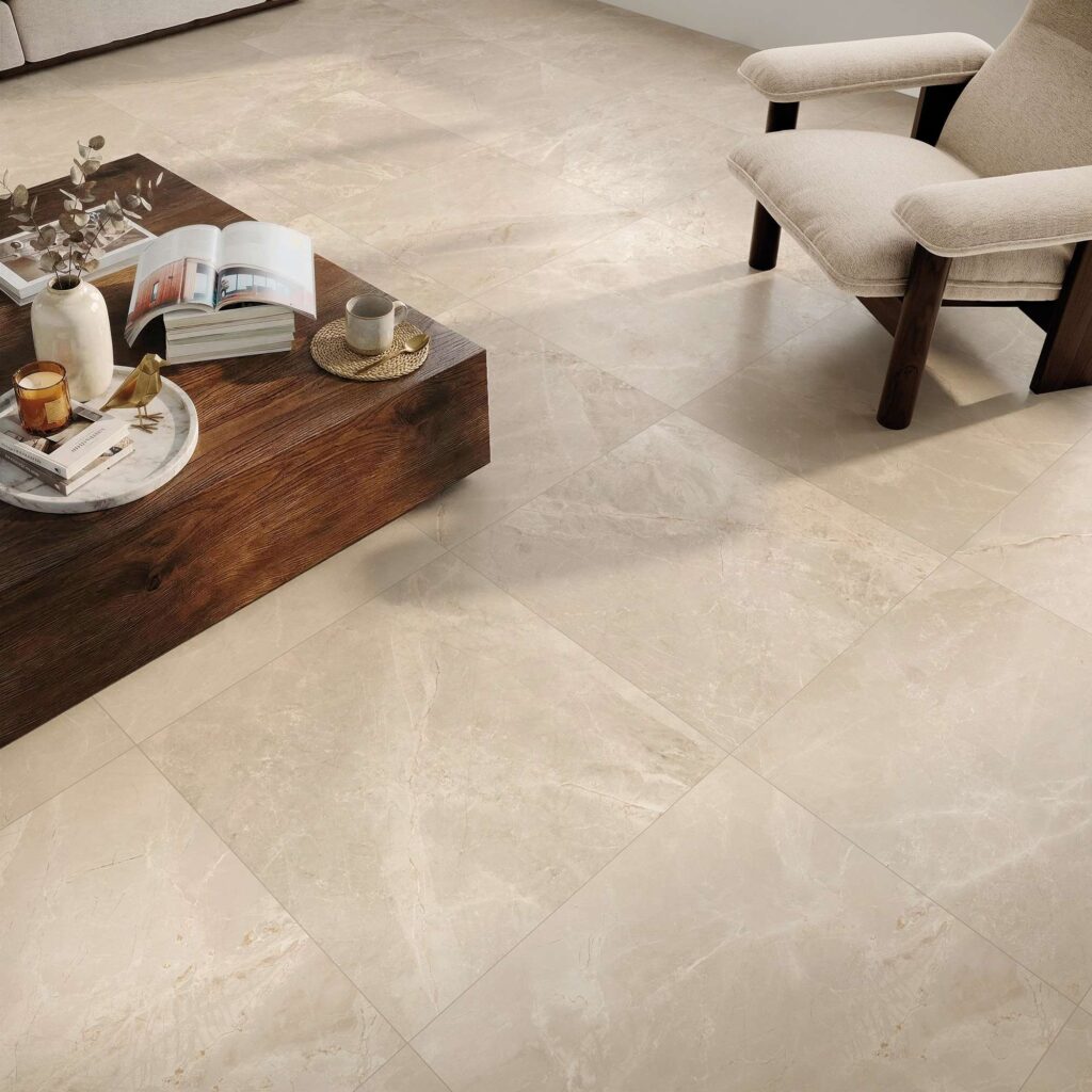 La collezione Elysion, nella variante colore Sand in formato 60x60, valorizza il pavimento di un moderno e luminoso soggiorno attraverso lastre in gres porcellanato effetto pietra dalle calde sfumature beige. La posa ortogonale esalta le delicate venature naturali della superficie, su cui poggiano un tavolino basso in legno scuro finemente decorato e una raffinata poltrona in tessuto chiaro, creando un'atmosfera accogliente e di sobria eleganza.