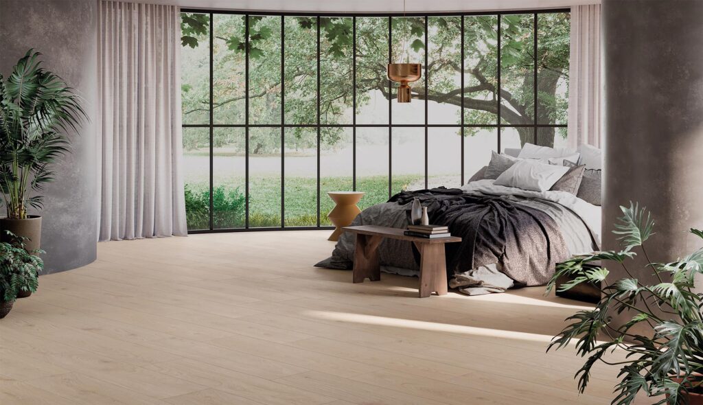 La collezione Barrique presenta un pavimento in gres porcellanato effetto legno Almond Natural nel formato 30x120 cm, posato a correre all'interno di un'ampia e luminosa camera da letto di design. La stanza, caratterizzata da un'atmosfera calda e rilassante, è valorizzata da una grande vetrata curva che si affaccia su un giardino alberato, inondando lo spazio di luce naturale. L'arredo elegante include un letto con tessuti nei toni del grigio e del bianco, una panca in legno rustico ai piedi del letto e rigogliose piante ornamentali che completano l'estetica naturale dell'ambiente.