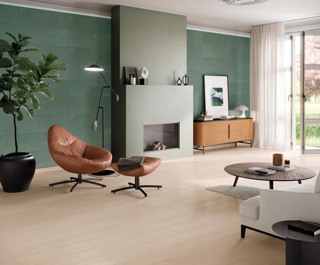 Pavimento in gres porcellanato effetto legno della collezione Barrique, nella tonalità chiara Almond e formato 20x120 cm, posato all'interno di un elegante e luminoso ambiente living. La superficie ceramica dal design naturale si estende in un ampio soggiorno moderno, caratterizzato da una parete di fondo color verde salvia con camino incassato, una raffinata poltrona di design in pelle marrone con pouf coordinato, un divano chiaro e dettagli in legno. La luce naturale, proveniente da un'ampia vetrata sullo sfondo, esalta le venature calde e la texture realistica del pavimento.