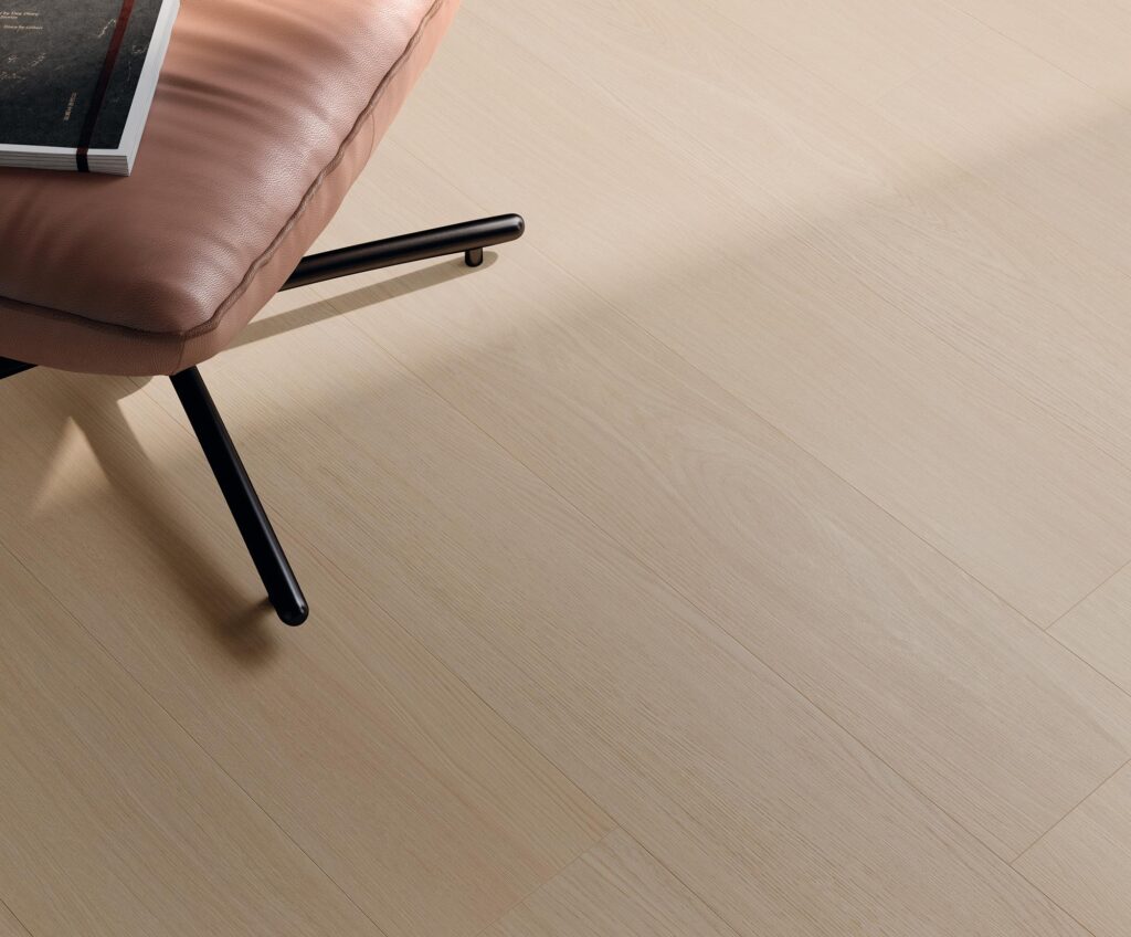 La collezione BARRIQUE in gres porcellanato effetto legno color Almond, nel formato 20x120, definisce lo stile di un elegante soggiorno contemporaneo. Il dettaglio del pavimento mostra una posa a correre con calde sfumature beige chiare e venature naturali, accostato a una raffinata sedia di design in pelle rosa cipria con gambe nere, evocando un'atmosfera accogliente, luminosa e minimalista.