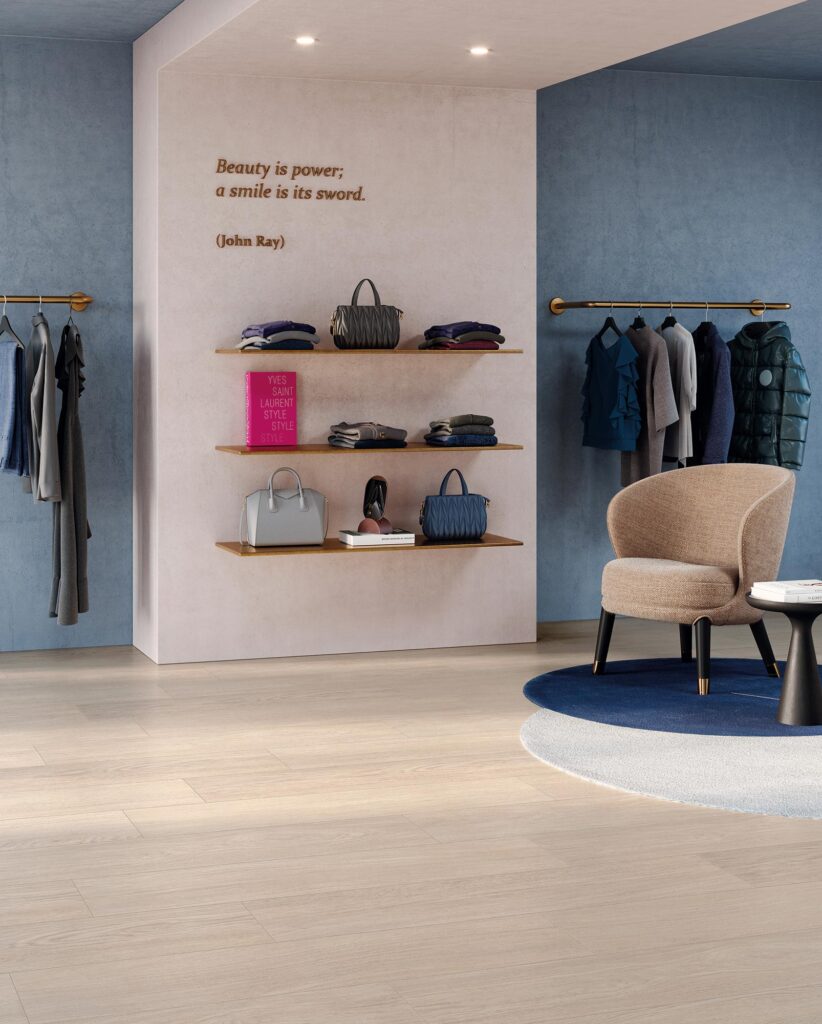 La collezione Barrique, nel caldo colore Almond e formato 20x120, riveste con eleganza il pavimento di una boutique di moda raffinata. Le doghe in gres porcellanato effetto legno sono posate longitudinalmente a correre, donando un senso di ampiezza e luminosità naturale allo spazio commerciale. L'ambiente di design presenta pareti azzurro polvere che contrastano delicatamente con un pilastro centrale panna, attrezzato con mensole dorate che espongono borse e abiti. Completa la scena un'area relax accogliente con una poltroncina beige, un tavolino nero e un tappeto circolare bicolore blu e grigio.