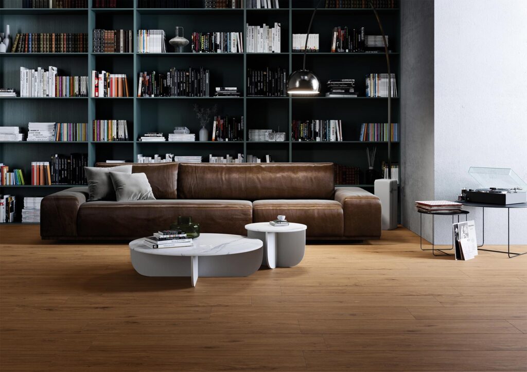 La collezione BARRIQUE veste questo elegante soggiorno con il gres porcellanato effetto legno nella variante Cherry Natural, formato 20x120 cm. Il pavimento, caratterizzato da una posa a correre che esalta le sfumature calde e naturali del ciliegio, si inserisce in un ambiente raffinato e intellettuale, definito da una grande libreria a tutta parete verde scuro, un divano in pelle marrone, tavolini di design con piano in marmo e un'illuminazione d'atmosfera.