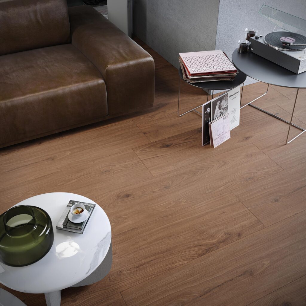La collezione Barrique propone un pavimento in gres porcellanato effetto legno nella calda tonalità Cherry Natural, formato 20x120 cm, caratterizzato da una posa sfalsata che esalta le venature e i dettagli naturali del legno. L'ambientazione, un angolo soggiorno intimo e ricercato, è arricchita da un arredamento elegante che comprende un divano in pelle marrone, un tavolino tondo in marmo bianco con una tazzina da caffè in primo piano e una zona d'ascolto retro con giradischi e dischi in vinile su tavolini in metallo nero.