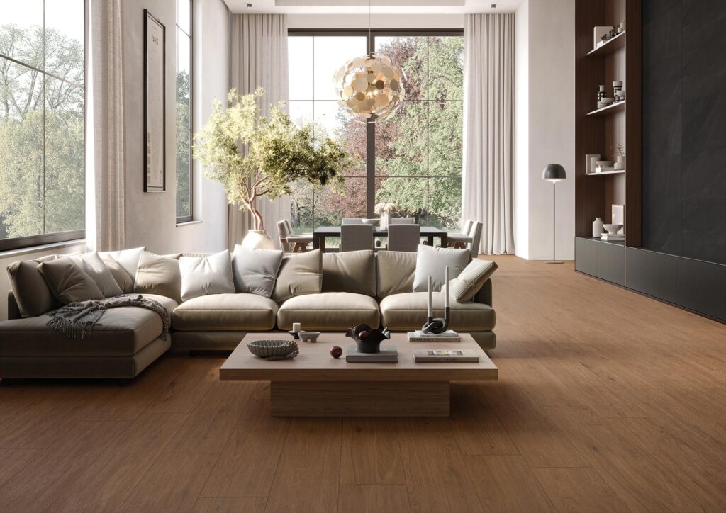La collezione di gres porcellanato effetto legno BARRIQUE, nella calda tonalità Cherry Natural e formato 20x120, definisce lo stile di un elegante e luminoso ambiente living. L'immagine mostra un ampio soggiorno di design contemporaneo con un grande divano ad angolo in tessuto beige, un tavolino da fumo centrale e spettacolari vetrate a tutta altezza aperte sul verde esterno. La posa a correre delle doghe effetto legno enfatizza la profondità dello spazio, creando un'atmosfera calda, accogliente e di raffinata armonia.