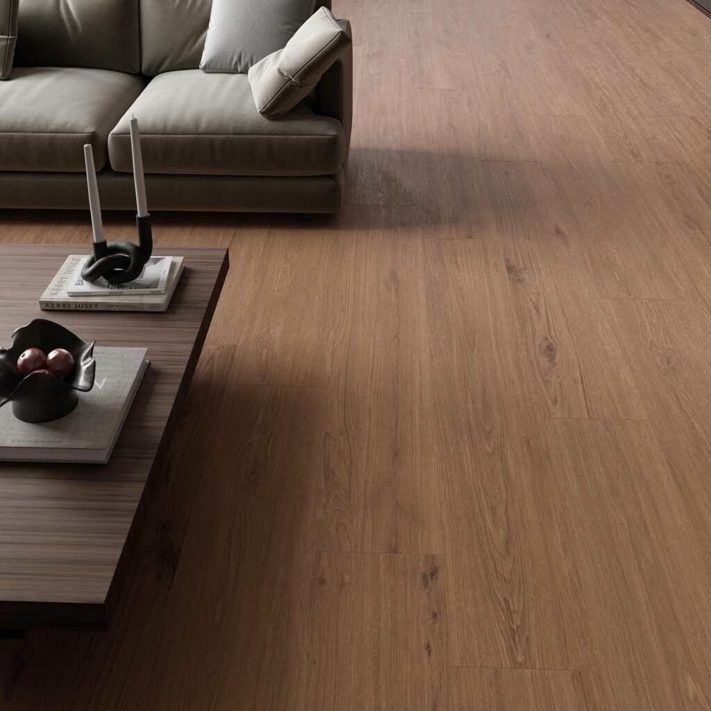 La collezione BARRIQUE in gres porcellanato effetto legno, nella calda tonalità Cherry Natural e formato 20x120 cm, riveste il pavimento di un elegante e accogliente soggiorno moderno. Le doghe, posate in modo sfalsato a correre, mettono in risalto la texture naturale con venature e nodi realistici ispirati al legno di ciliegio. L'ambiente living è completato da un divano di design nei toni del grigio e da un tavolino da caffè in legno scuro con libri e oggetti decorativi raffinati, che valorizzano l'atmosfera intima e contemporanea.