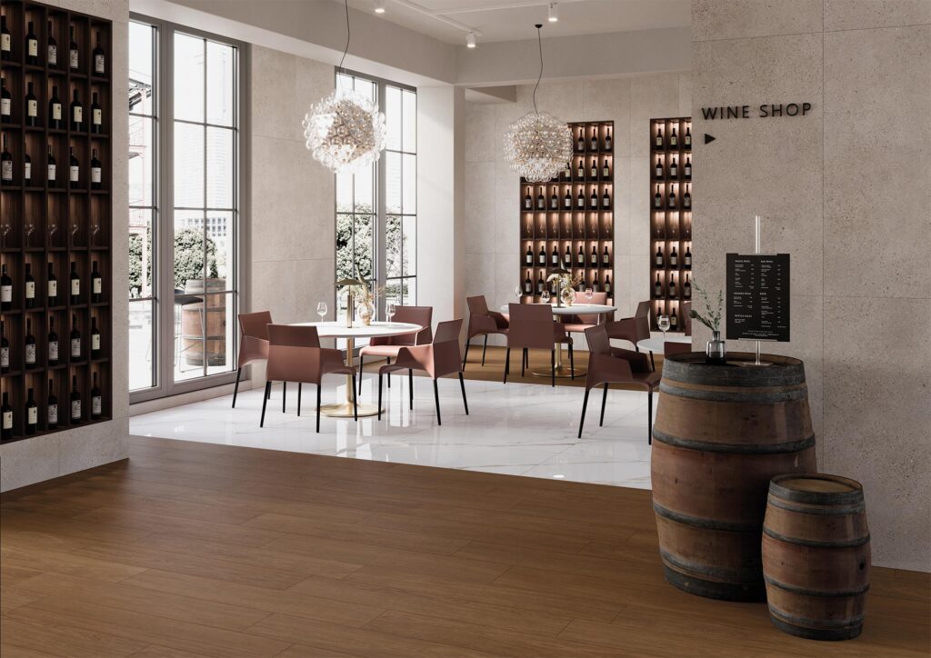 La collezione BARRIQUE di piastrelle in gres porcellanato effetto legno, nella calda tonalità Cherry Selected formato 20x120, caratterizza con una posa a correre il pavimento di un elegante ristorante con wine shop. L'ambiente, moderno e luminoso, mostra in primo piano le doghe ceramiche che richiamano il legno naturale, accostate a botti decorative; sullo sfondo, la zona pranzo presenta un raffinato contrasto con un pavimento in lastre chiare effetto marmo lucido, completata da tavoli rotondi, poltroncine dal design contemporaneo e scaffalature per il vino integrate nelle pareti chiare.