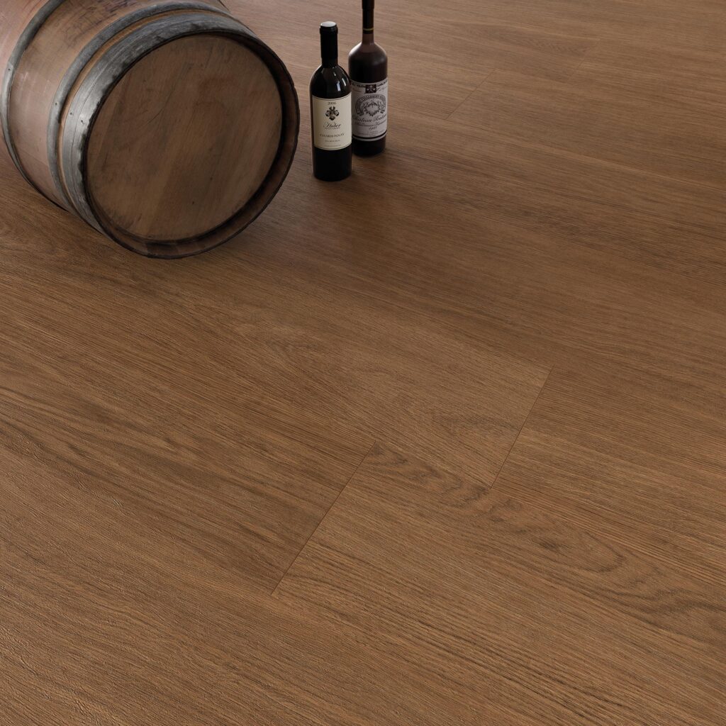 Collezione BARRIQUE in gres porcellanato effetto legno nella calda variante Cherry, formato 20x120. Il pavimento, caratterizzato da una posa sfalsata a correre che valorizza le venature naturali del legno, è presentato in un'ambientazione per ristorante dall'atmosfera calda e accogliente, evocata dalla presenza di una botte in legno e due bottiglie di vino adagiate sulla superficie.