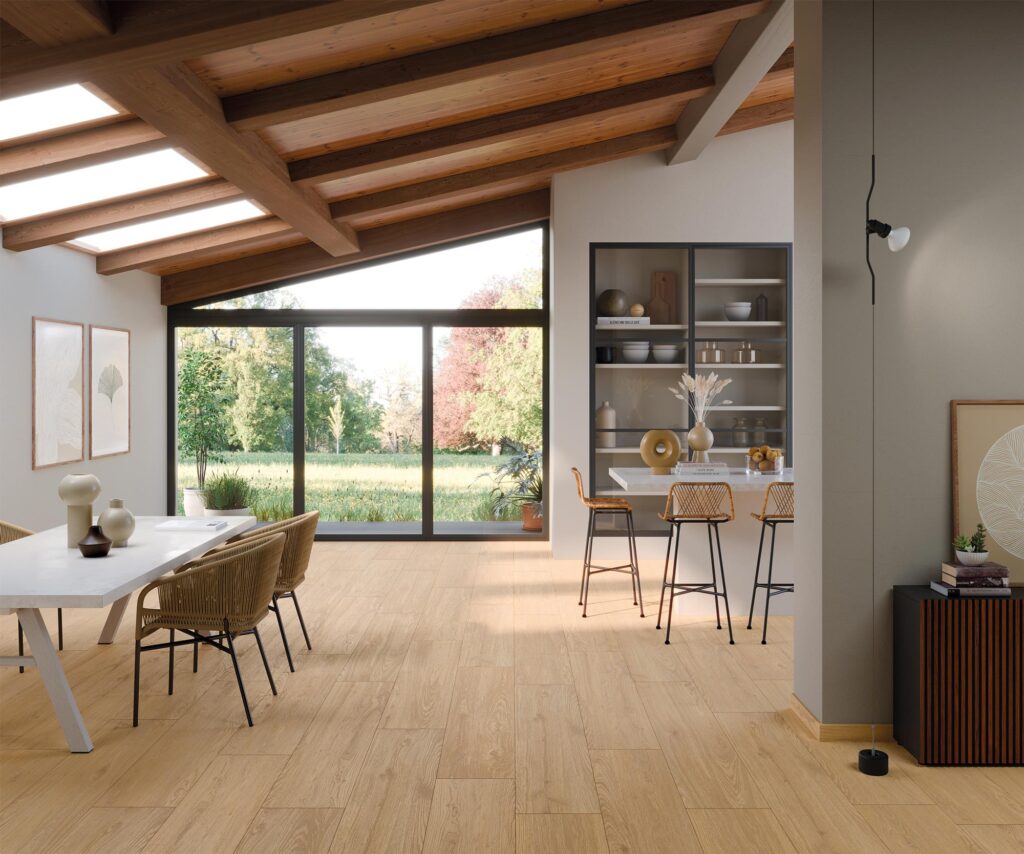 La collezione BARRIQUE presenta un pavimento in gres porcellanato effetto legno nella tonalità Honey Natural, formato 30x120 cm, posato a correre in un prestigioso e luminoso ambiente living open space. L'immagine mostra un soggiorno di design con soffitto spiovente caratterizzato da travi in legno a vista e ampie vetrate affacciate su un giardino verdeggiante. L'arredamento coordinato, con tavolo da pranzo e isola bar dai dettagli in paglia e legno, esalta l'atmosfera calda, naturale ed elegante creata dalle venature e dai nodi realistici delle doghe ceramiche.