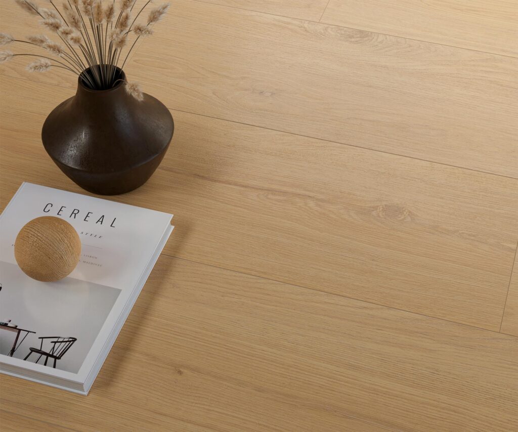 La collezione Barrique presenta il gres porcellanato effetto legno Honey Natural nel formato 30x120 cm, ideale per una zona living calda e raffinata. L'immagine mostra un dettaglio ravvicinato della pavimentazione con posa a listoni, di cui si apprezzano le venature realistiche e le morbide sfumature color miele. In un angolo, una composizione decorativa con un vaso scuro contenente rami secchi, una rivista di design e una sfera in legno esalta l'atmosfera minimalista, accogliente ed elegante dell'ambiente.