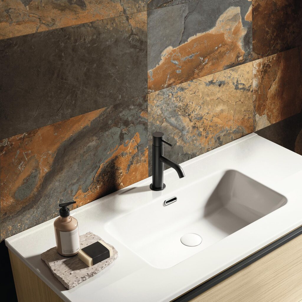 La collezione BET, nella suggestiva variante Indian Summer in formato 30x60 cm, riveste la parete di un bagno dal design moderno e raffinato. L'inquadratura ravvicinata mostra un lavabo bianco con miscelatore nero opaco, accostato a piastrelle effetto ardesia posate orizzontalmente, caratterizzate da un affascinante contrasto cromatico tra il grigio scuro e calde venature color ruggine e rame.