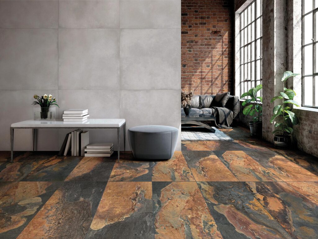 La collezione BET, nella variante colore Indian Summer in formato 60x120 cm, caratterizza il pavimento di questo spazioso e luminoso soggiorno in stile loft industriale. Le piastrelle in gres porcellanato effetto pietra, ricche di calde sfumature ruggine, rame e grigio antracite, sono posate a correre per donare dinamismo e profondità all'ambiente. Sulla sinistra, una parete grigia minimalista fa da sfondo a un tavolino bianco con libri e a un pouf grigio. Sullo sfondo, la zona living accoglie un elegante divano in pelle nera davanti a una parete con mattoni rossi a vista, illuminata da ampie finestre industriali decorate con piante verdi in vaso.