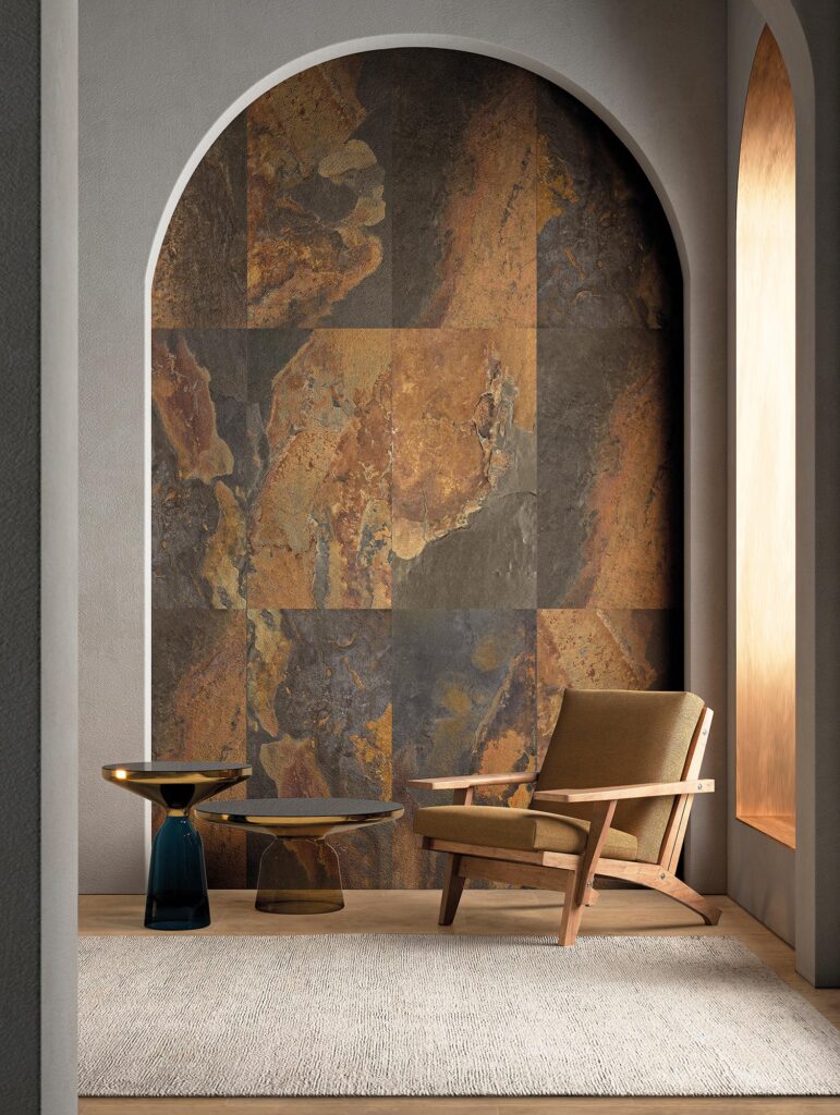 La collezione BET, nella variante cromatica Indian Summer formato 60x60, valorizza una scenografica parete ad arco in gres porcellanato effetto pietra con calde venature ruggine, antracite e dorate. L'immagine mostra un raffinato angolo lounge dal design contemporaneo, dove il rivestimento materico fa da sfondo a un'elegante poltrona in legno con seduta color senape, affiancata da due tavolini da caffè in vetro e metallo lucido su un tappeto chiaro, infondendo all'ambiente un'atmosfera lussuosa e accogliente.