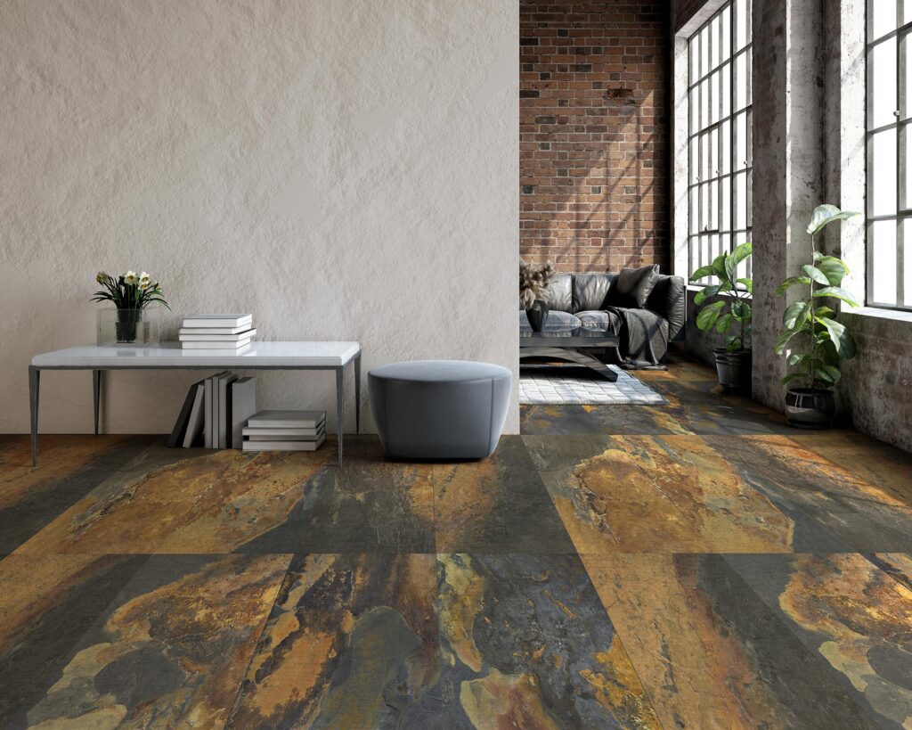 La collezione BET, nella variante cromatica Indian Summer in formato 60x60, valorizza questo moderno e luminoso soggiorno in stile industrial chic. Il pavimento in gres porcellanato presenta una texture materica ricca di sfumature calde color ruggine, ocra e grigio scuro, che richiamano la pietra naturale. Sulla sinistra, una parete chiara e texturizzata fa da sfondo a un tavolino minimalista con libri e a un pouf grigio, mentre sullo sfondo si apre una spaziosa zona living caratterizzata da una parete con mattoni rossi a vista, grandi finestre di stampo industriale, un divano in pelle nera e rigogliose piante d'appartamento.