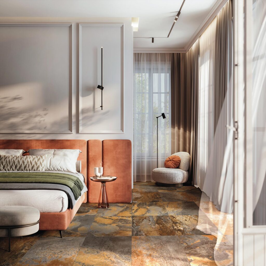 La collezione BET, nella variante cromatica Indian Summer in formato 60x60, definisce il carattere di questo raffinato ambiente grazie a un pavimento in gres porcellanato effetto pietra dalle calde sfumature ruggine, oro e grigio antracite. La posa ortogonale e regolare valorizza la texture naturale e materica delle piastrelle, che si sposa armoniosamente con l'elegante arredamento della stanza: un letto con testata in velluto color terracotta, pareti bianche decorate con boiserie e un angolo lettura minimalista con poltrona bouclé. La luce naturale, che filtra dalle ampie finestre velate da tende leggere, crea un'atmosfera calda, accogliente e sofisticata.