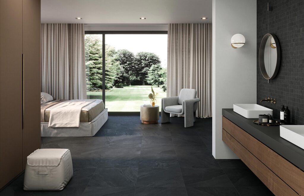 La collezione BET, nella variante colore Pyrite e formato 60x60, definisce il carattere di questa lussuosa camera da letto open space con bagno en-suite. Il pavimento in gres porcellanato scuro, con una texture effetto pietra naturale, dona profondità a un ambiente luminoso e raffinato, aperto verso un rigoglioso giardino esterno grazie a una grande vetrata scorrevole. Sulla sinistra si trova la zona notte con letto matrimoniale in tonalità neutre e una poltrona moderna; sulla destra, l'elegante zona bagno presenta un mobile sospeso in legno con lavabi da appoggio bianchi, esaltati da una parete di fondo rivestita con un delicato mosaico coordinato che completa l'atmosfera contemporanea e di design.