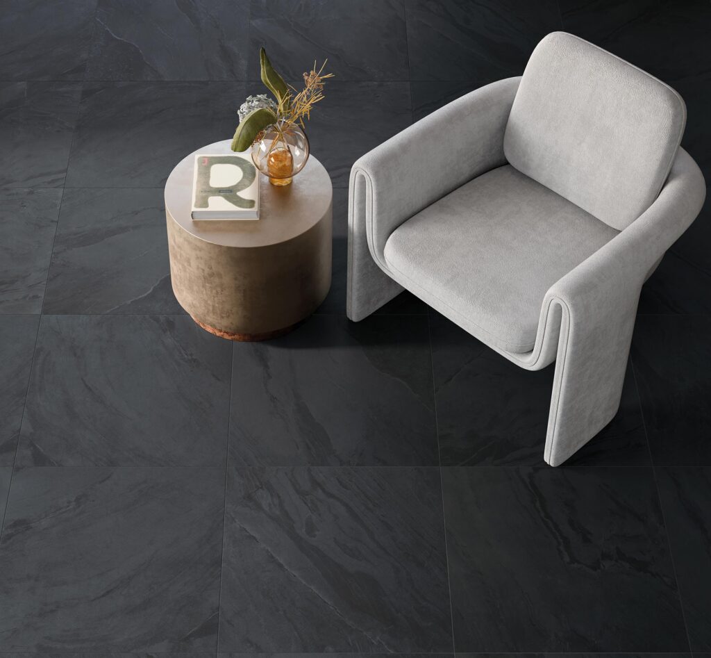 La collezione BET, nella variante colore Pyrite e formato 60x60 cm, definisce con eleganza il pavimento di un raffinato ambiente bedroom. La vista dall'alto mette in risalto la texture materica e naturale delle piastrelle in gres porcellanato effetto pietra scura, posate in modo regolare. Su questo sfondo scuro e profondo spiccano una moderna poltrona di design in tessuto grigio chiaro e un tavolino cilindrico in metallo brunito, impreziosito da un libro e un piccolo vaso con elementi floreali secchi.