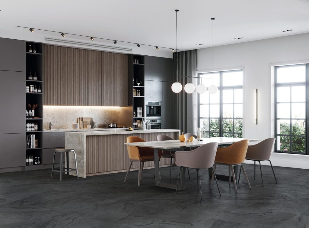 La collezione BET in gres porcellanato, qui presentata nella tonalità scura Pyrite nel formato 60x60, definisce con eleganza il pavimento di una cucina moderna e di design con posa regolare. Le piastrelle effetto pietra, caratterizzate da una texture materica e venature naturali, creano un sofisticato contrasto con l'isola centrale rivestita in legno e marmo chiaro. La scena si apre su una luminosa zona pranzo con tavolo coordinato e poltroncine avvolgenti nei toni caldi dell'ocra e del tortora, inserite in un ambiente raffinato illuminato da grandi vetrate e luci di design sospese.