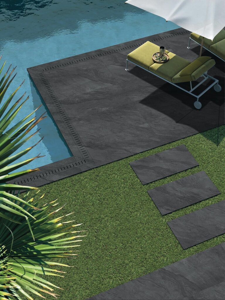 La collezione BET nell'intensa tonalità Pyrite veste uno spazio esterno di design con lastre in gres porcellanato nel formato 60x90 cm e spessore 2 cm. La veduta dall'alto mostra una raffinata zona piscina con pavimentazione scura effetto pietra naturale, completata da un camminamento con posa a secco su prato verde, eleganti lettini prendisole e un ombrellone, evocando un'atmosfera lussuosa e rilassante.