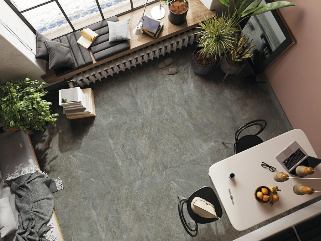 La collezione BET, proposta nella variante colore Silver Gray nel formato 60x60 cm, valorizza un elegante e luminoso ambiente living in questa raffinata vista dall'alto. Il pavimento in gres porcellanato effetto pietra grigia, caratterizzato da una posa regolare e venature naturali, fa da sfondo a un arredamento moderno composto da un tavolo bianco con sedie nere, piante verdi rigogliose e una confortevole seduta sotto la finestra arricchita da cuscini e libri, per un'atmosfera contemporanea e accogliente.