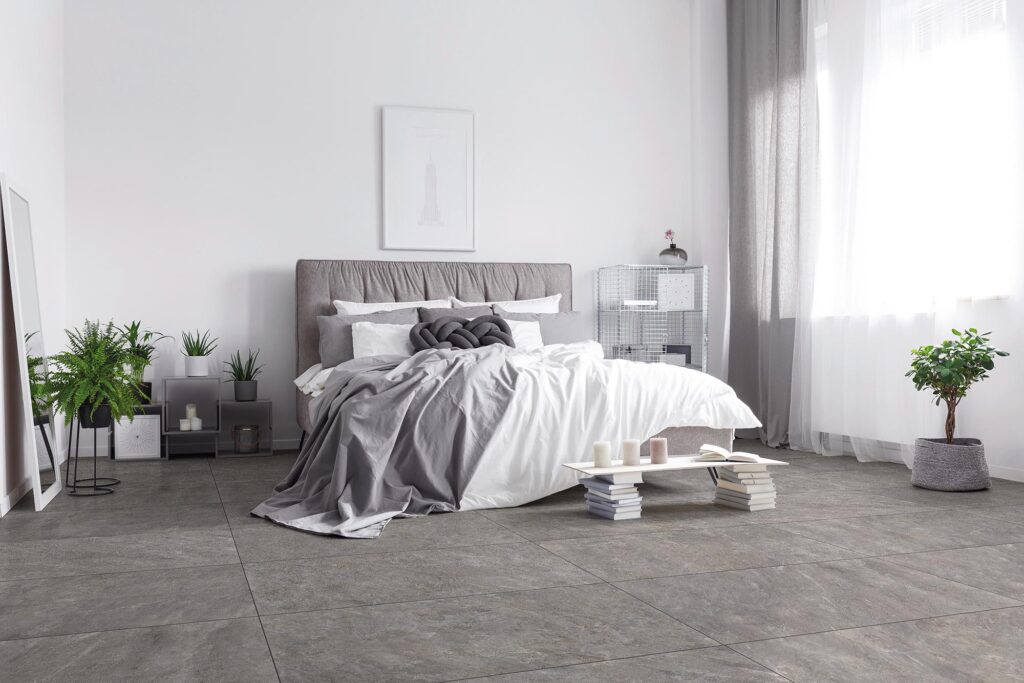 Pavimento in gres porcellanato della collezione BET nel colore Silver Grey e formato 60x120 cm, posato in modo regolare in un'elegante e luminosa camera da letto moderna. L'ambiente, caratterizzato da un'atmosfera minimalista e rilassante, presenta un letto matrimoniale con testiera e tessili coordinati nei toni del grigio e del bianco, piante verdi in vaso, dettagli d'arredo essenziali e una grande finestra con tende leggere che illumina la texture materica effetto pietra delle piastrelle.