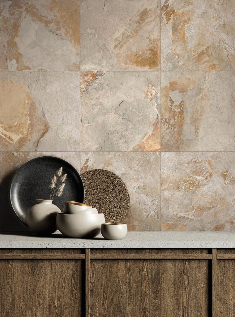 La collezione BET, nella variante Tomor in formato 60x60 cm, definisce questo raffinato rivestimento a parete in gres porcellanato effetto pietra, caratterizzato da intense sfumature calde che sfumano dal grigio terra all'ocra e al ruggine. In primo piano, su una credenza in legno scuro con top chiaro, è disposta una composizione decorativa di stoviglie di design in ceramica beige e oro, un piatto scuro e spighe secche, che esalta la texture materica delle piastrelle creando un'atmosfera intima, calda e sofisticata.