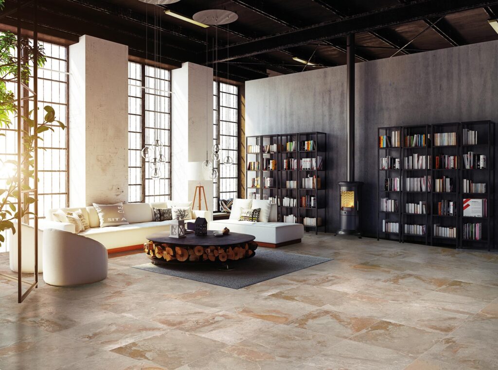 La collezione BET, nella variante TOMOR in formato 60x60 cm, definisce con eleganza il pavimento di un ampio living in stile loft industriale. Le piastrelle in gres porcellanato effetto pietra naturale mostrano una ricca stonalizzazione che sfuma dal beige caldo alle tonalità del grigio e del ruggine. L'ambiente, inondato dalla luce naturale che filtra da grandi finestre a riquadri, è arredato con divani moderni di colore chiaro, un originale tavolino tondo con legna a vista, librerie in metallo nero a tutta altezza e una stufa di design accesa, creando un'atmosfera accogliente e sofisticata.