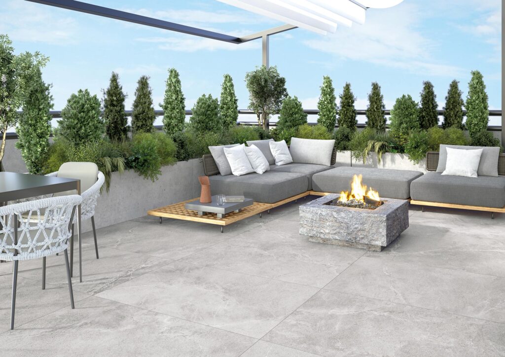 La collezione BLEND veste questo elegante spazio outdoor con un pavimento in gres porcellanato color Ash nel formato 60x120 cm. L'ampio terrazzo dal design moderno è allestito con un raffinato divano angolare grigio e cuscini bianchi, un suggestivo braciere in pietra con fiamma accesa e fioriere perimetrali ricche di arbusti verdi, creando un'atmosfera lussuosa e accogliente ideale per il relax all'aperto.