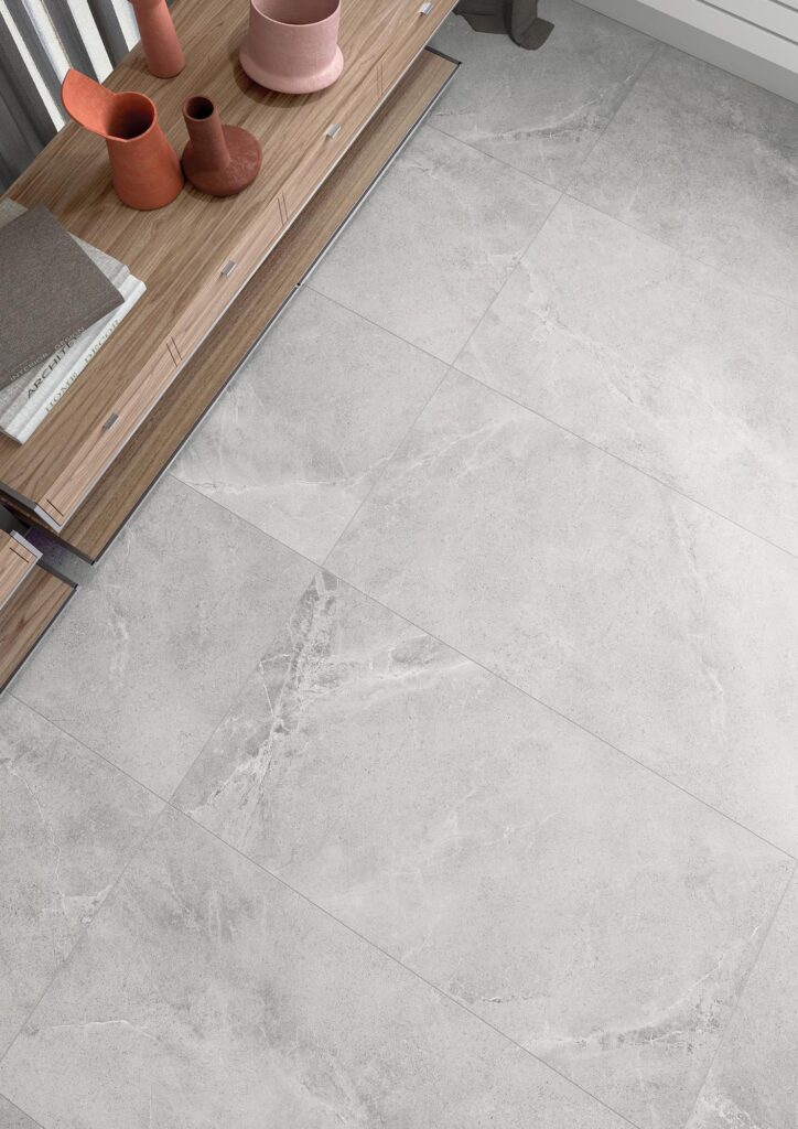 La collezione BLEND, qui presentata nella tonalità grigia Ash e nel formato 60x120 cm, riveste il pavimento di un moderno ambiente living. L'immagine mostra una raffinata posa di piastrelle in gres porcellanato effetto pietra con delicate venature chiare. Sulla sinistra, un mobile in legno ospita vasi decorativi color terracotta e libri di design, creando un'atmosfera elegante e accogliente.