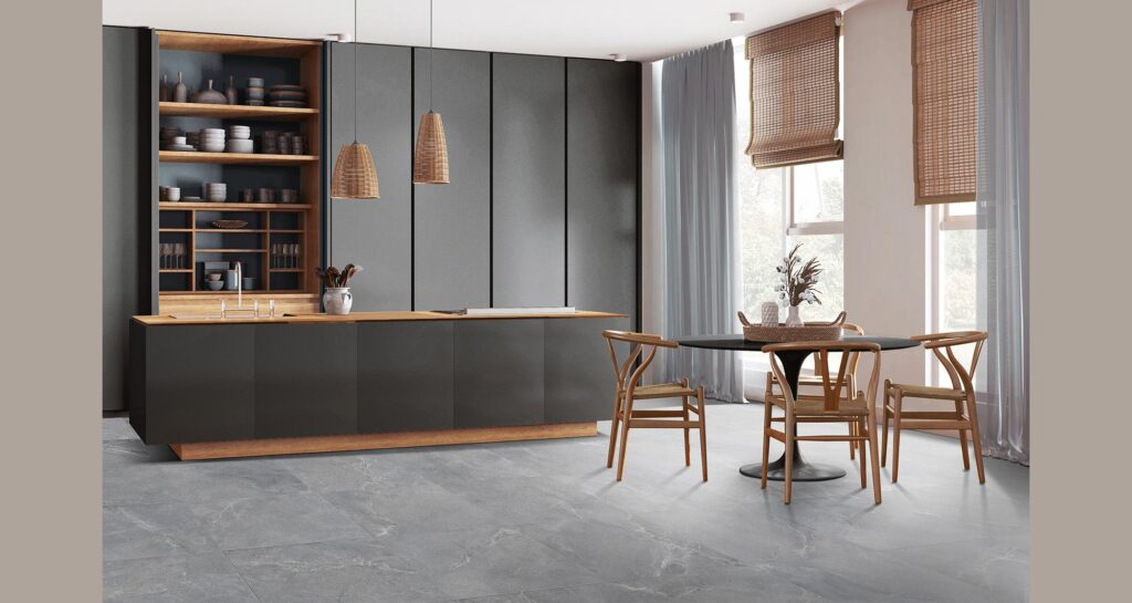 Collezione BLEND, variante colore Ash nel formato 60x60 cm. Un elegante pavimento in gres porcellanato effetto pietra grigia, posato in modo regolare, definisce lo spazio di una cucina moderna, luminosa e di design. L'ambiente è caratterizzato da un'isola scura con top in legno caldo, una raffinata zona pranzo con tavolo rotondo nero e sedie in legno chiaro stile Wishbone, il tutto inondato dalla luce naturale che filtra da ampie finestre con tende e stuoie in bambù.