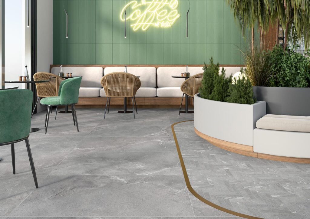 La collezione BLEND veste il pavimento di un moderno e luminoso bar di design con grandi lastre in gres porcellanato effetto pietra nella tonalità grigia Rock, formato 60x120. Sulla destra, un profilo metallico dorato delimita un'elegante area decorativa definita dalla posa a spina di pesce del mosaico Arrow. L'ambiente, fresco e accogliente, è arredato con tavolini da bistrot, poltroncine in velluto verde e in fibra naturale, una panca coordinata a parete e grandi fioriere con piante verdi. Sullo sfondo spicca una parete verde salvia impreziosita da una scritta al neon gialla "Coffee".