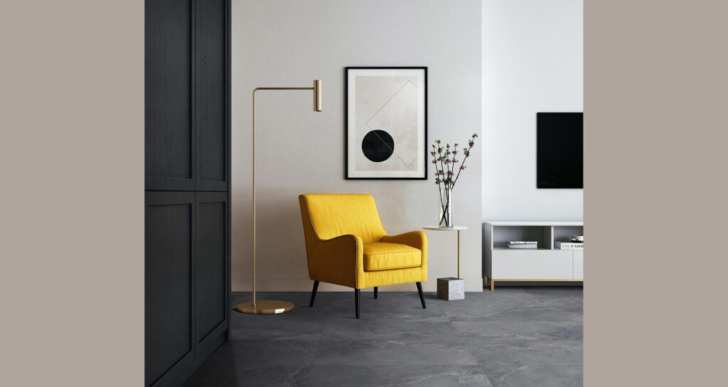 La collezione Blend, nella variante Rock in formato 60x120 cm, definisce il pavimento di questo raffinato ambiente living dal design contemporaneo. Le grandi lastre in gres porcellanato effetto pietra grigio scuro creano una superficie materica e continua, su cui spicca in forte contrasto una poltrona di un vivace giallo senape, affiancata da un'elegante lampada da terra dorata e da un tavolino minimalista. Sullo sfondo, una parete grigio chiaro ospita un quadro geometrico astratto, completando un'atmosfera luminosa, sofisticata e di design.