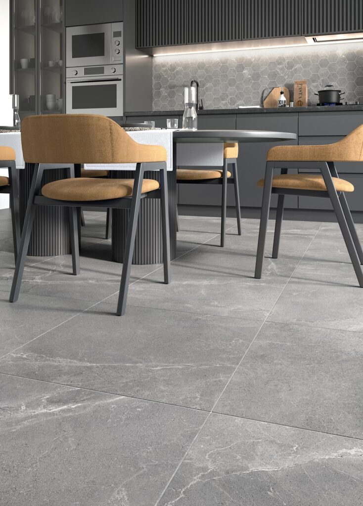 La collezione BLEND, nella variante Rock effetto pietra grigia in formato 60x60, valorizza il pavimento di una moderna cucina di design. L'inquadratura in primo piano mostra la posa regolare delle piastrelle in gres porcellanato, caratterizzate da venature naturali e dettagli materici. Sullo sfondo, un elegante tavolo da pranzo con sedie imbottite color senape dialoga con i mobili della cucina grigio scuro e un raffinato paraspruzzi a mosaico esagonale retroilluminato, creando un'atmosfera calda e contemporanea.