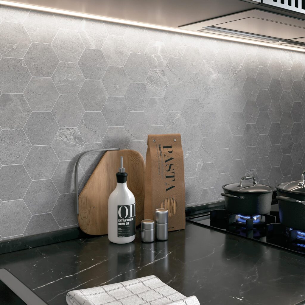 La collezione Blend, nella variante colore Rock, riveste con eleganza la parete di una cucina moderna attraverso un raffinato mosaico esagonale grigio effetto pietra. L'immagine in primo piano mostra il paraschizzi valorizzato da una luce LED sottopensile che esalta la texture materica e naturale delle tessere. Sul piano di lavoro in marmo nero venato sono accostati un tagliere in legno, una bottiglia di olio d'oliva, un pacchetto di pasta e alcune pentole sui fuochi accesi, ricreando un'atmosfera calda, accogliente e di design.