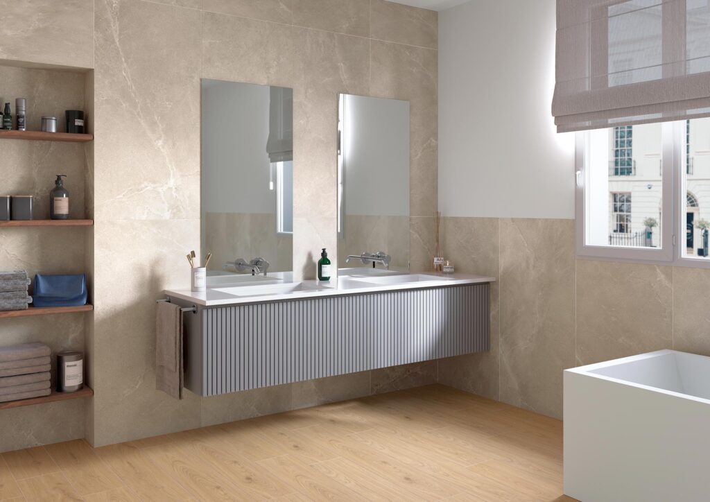 La collezione BLEND, qui nella calda e sofisticata tonalità Tan in formato 60x120 cm, riveste con eleganza le pareti di un bagno moderno e luminoso. Le grandi lastre in gres porcellanato effetto pietra sono posate a tutta altezza, creando una superficie continua e raffinata che fa da sfondo a un mobile lavabo sospeso grigio con texture cannettata e doppio specchio. L'ambiente, caratterizzato da un'atmosfera serena e accogliente, è completato da una nicchia a muro con mensole in legno, una moderna vasca da bagno bianca e un pavimento in gres effetto legno che scalda l'intera stanza.