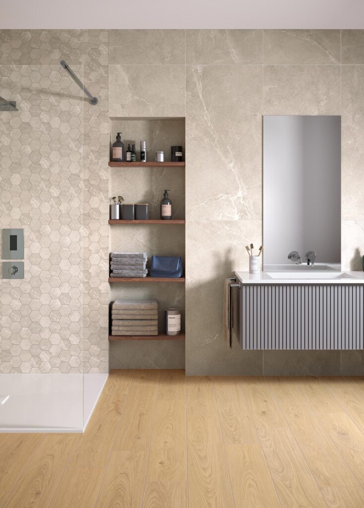 La collezione BLEND, nella variante cromatica Tan in formato 60x120 cm, definisce l'atmosfera calda e raffinata di questo bagno contemporaneo. La parete principale presenta una posa verticale di grandi lastre effetto pietra beige arricchite da delicate venature, accostate a una zona doccia rivestita con un elegante mosaico esagonale coordinato. Al centro, una nicchia incassata con mensole in legno ospita asciugamani e accessori, mentre sulla destra spicca un mobile bagno sospeso grigio con texture cannettata e lavabo integrato, sormontato da uno specchio rettangolare. Il pavimento in gres porcellanato effetto legno chiaro completa l'ambiente infondendo armonia e luminosità.