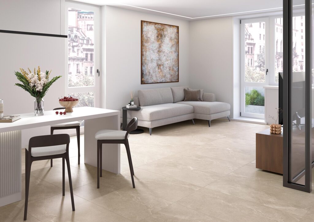 La collezione BLEND in gres porcellanato color Tan, nel formato 60x120 cm, riveste con eleganza il pavimento di un moderno e luminoso ambiente living. L'immagine mostra la posa di grandi lastre ceramiche dalle calde tonalità beige effetto pietra naturale, che donano ampiezza e continuità visiva allo spazio. La stanza, caratterizzata da un'atmosfera accogliente e raffinata, è arredata in stile contemporaneo con un divano angolare grigio chiaro, un'isola bianca con sgabelli di design e grandi vetrate che inondano la zona giorno di luce naturale.