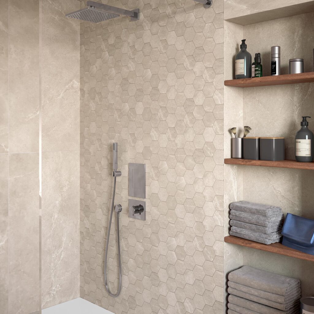Collezione BLEND: rivestimento per bagno nell'elegante tonalità Tan con formato mosaico esagonale. L'immagine mostra un raffinato primo piano di un moderno ambiente doccia con rubinetteria cromata, caratterizzato da una parete d'accento in piastrelle esagonali beige dall'effetto pietra naturale. Sulla destra, una nicchia integrata con mensole in legno accoglie asciugamani grigi ordinatamente piegati e flaconi di bellezza, ricreando un'atmosfera calda, minimalista e rilassante da spa domestica.