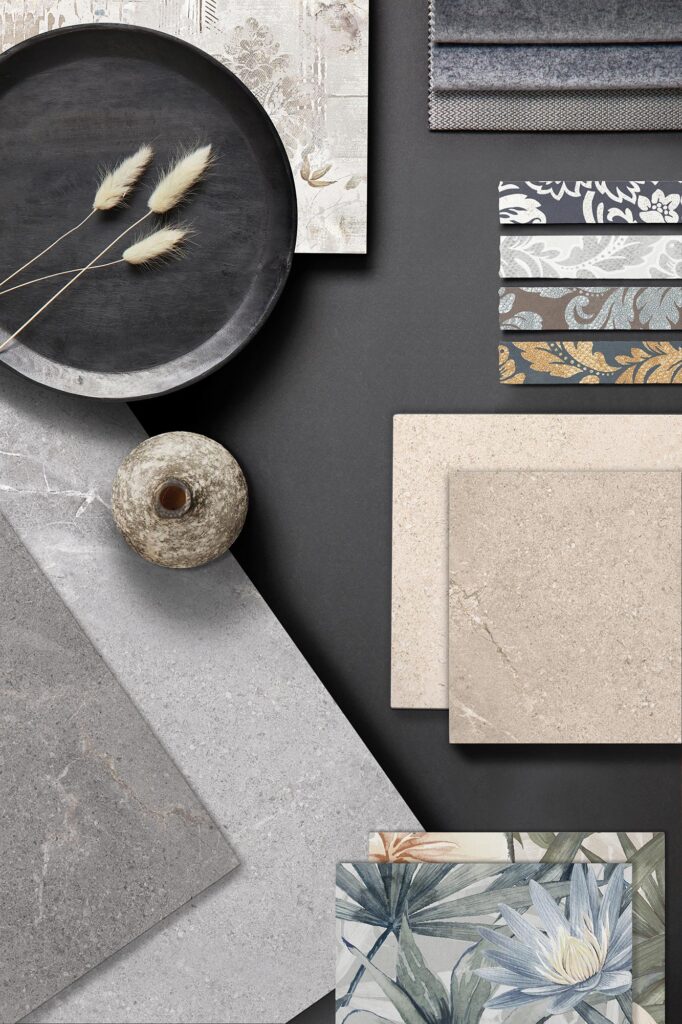 La collezione BLEND presentata in un'elegante moodboard d'ispirazione per l'interior design su sfondo antracite. La composizione mette in mostra campioni di piastrelle in gres porcellanato effetto pietra e cemento, declinati nelle calde tonalità del beige e del crema e nelle sfumature minimali del grigio chiaro e scuro. Completano la selezione listelli decorati con motivi damascati dai dettagli dorati, una piastrella con decoro botanico stile acquerello con fiori di loto e foglie tropicali, e una selezione di tessuti coordinati in velluto grigio. L'atmosfera materica e sofisticata è arricchita da elementi di design come un vassoio nero con spighe decorative e un piccolo vaso in ceramica grezza.