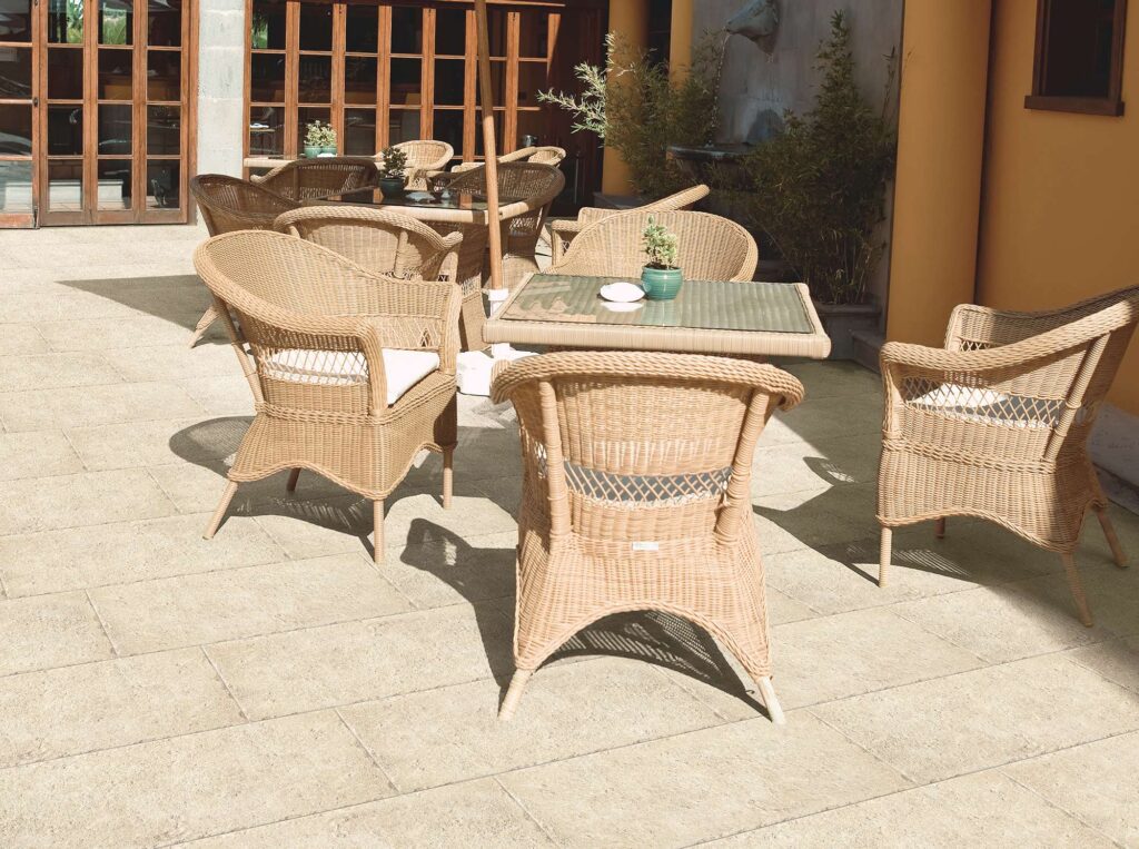 La collezione Bonnieux Beige, nel formato 30x60 per esterni, caratterizza la pavimentazione in gres porcellanato effetto pietra di un raffinato patio soleggiato. Le piastrelle, posate con schema sfalsato, mostrano una texture materica e naturale color beige che si integra armoniosamente con l'arredo outdoor composto da eleganti poltroncine e tavoli in rattan, piante decorative e ampie vetrate sullo sfondo, creando un'atmosfera accogliente e di classe.