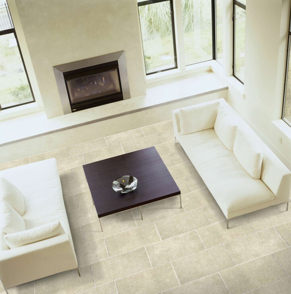La collezione Bonnieux Beige nel formato 30x60 cm definisce lo spazio di un raffinato soggiorno contemporaneo, mostrato in una suggestiva vista dall'alto. Il pavimento in gres porcellanato effetto pietra, con posa sfalsata a correre, esalta la texture naturale e la tonalità calda e luminosa della ceramica. L'ambiente living, elegante e minimalista, è arredato con due divani bianchi posizionati simmetricamente ai lati di un tavolino quadrato in legno scuro. Grandi vetrate e un moderno camino a parete diffondono una luce naturale che valorizza l'atmosfera spaziosa, accogliente e di design.
