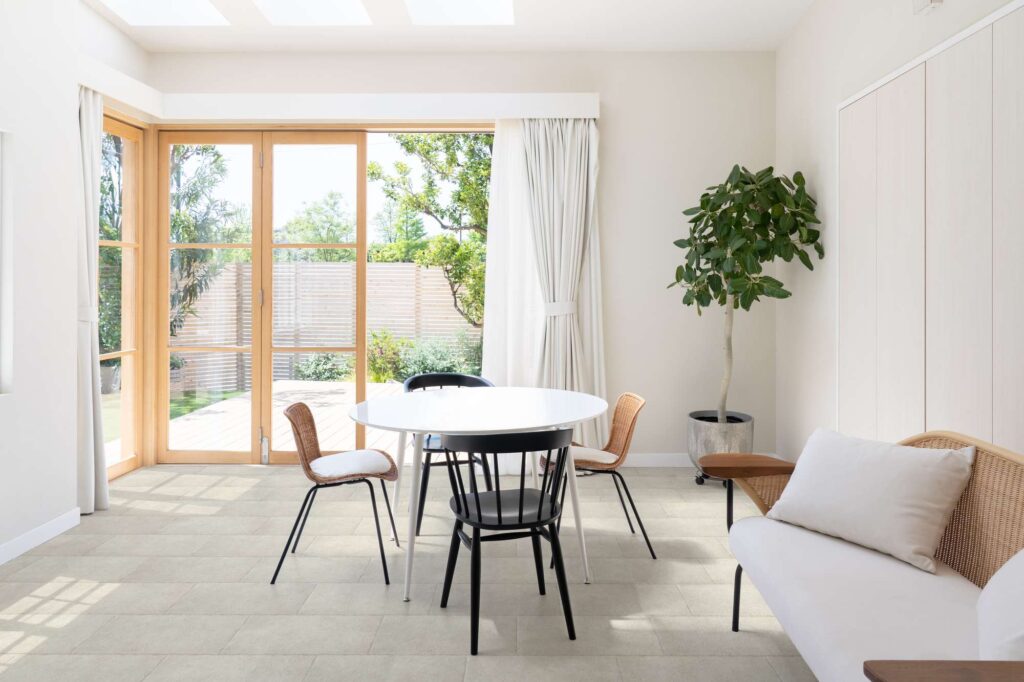 La collezione Bonnieux nella variante Beige, nel formato 30x60 cm, definisce con raffinatezza il pavimento di una luminosa zona giorno. Le piastrelle in gres effetto pietra, posate a correre (sfalsate), valorizzano un ambiente moderno e arioso arredato con un tavolo da pranzo rotondo bianco, sedie coordinate in legno nero e midollino, un divano di design chiaro e una rigogliosa pianta d'appartamento. Grandi vetrate in legno chiaro si affacciano su un giardino esterno, inondando lo spazio di luce naturale e sottolineando la texture naturale e la tonalità calda e accogliente della superficie ceramica.