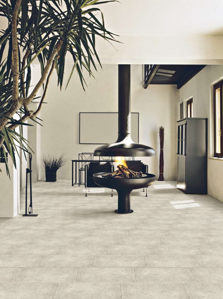 Collezione BONNIEUX PIERRE nel formato 30x60 cm: un elegante pavimento in gres porcellanato effetto pietra dalle tonalità chiare e naturali del beige, posato a correre (sfalsato) in un ampio e luminoso ambiente living. La scena, dall'atmosfera calda e accogliente, mostra al centro della stanza un moderno caminetto sospeso di design nero con fuoco acceso, circondato da arredi minimalisti scuri, una grande pianta verde in primo piano e pareti chiare che esaltano la texture materica e la raffinatezza delle superfici.