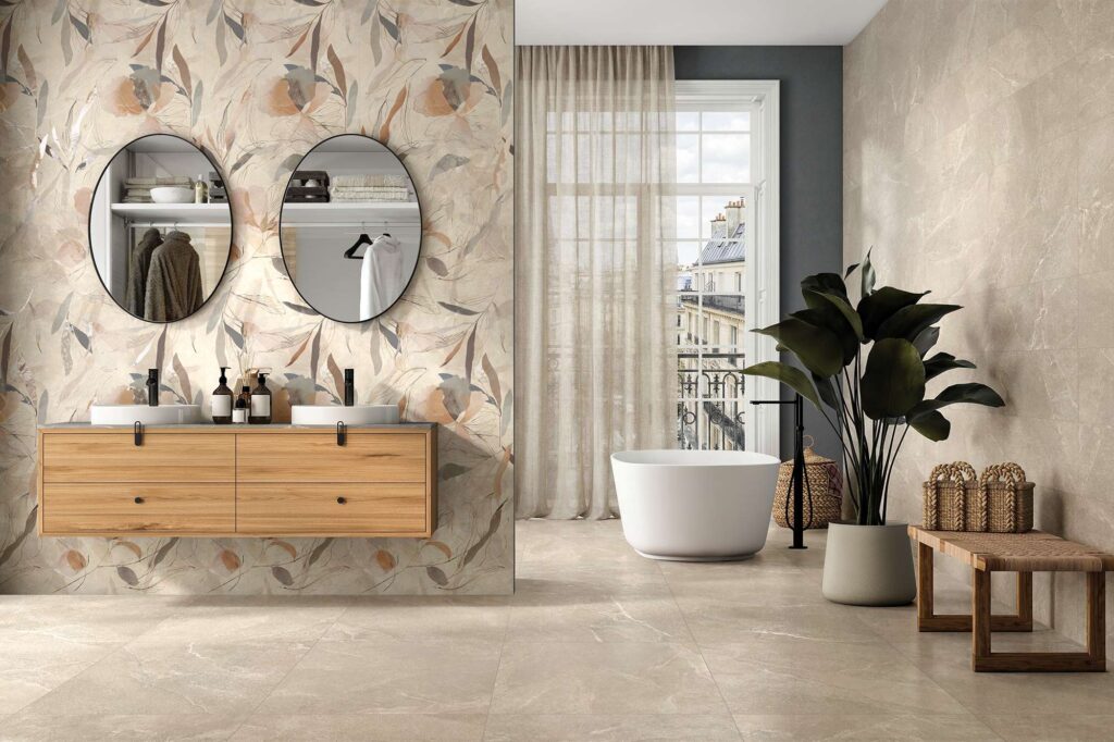 La collezione Reshape definisce un ambiente bagno raffinato e luminoso, proponendo le varianti Sand e il decoro Acquarelle nei formati 60x60, 30x60 e 60x120. L'immagine mostra una parete d'accento rivestita con il decoro floreale stile acquerello, su cui sono installati due specchi ovali neri e un elegante mobile lavabo sospeso in legno chiaro con doppi lavabi da appoggio. Il pavimento e la parete adiacente sono rivestiti con piastrelle effetto pietra nella tonalità sabbia calda. Sullo sfondo, una vasca freestanding bianca posizionata davanti a una grande finestra con tenda leggera infonde un senso di benessere e relax.