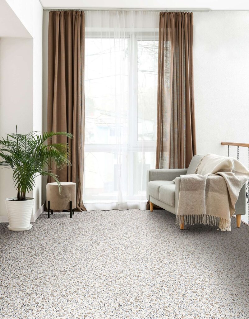 La collezione di piastrelle D-Lash nella calda variante colore Iced Mocha veste il pavimento di un luminoso soggiorno con un elegante effetto terrazzo alla veneziana. Le lastre ceramiche in formato quadrato, posate con fughe sottili, creano una superficie continua e armoniosa in perfetta sintonia con gli arredi circostanti: un divano grigio chiaro con una morbida coperta beige, una pianta d'appartamento in un vaso bianco e una grande finestra con doppie tende marroni e bianche che lasciano filtrare la luce naturale, donando all'ambiente un'atmosfera accogliente e rilassante.