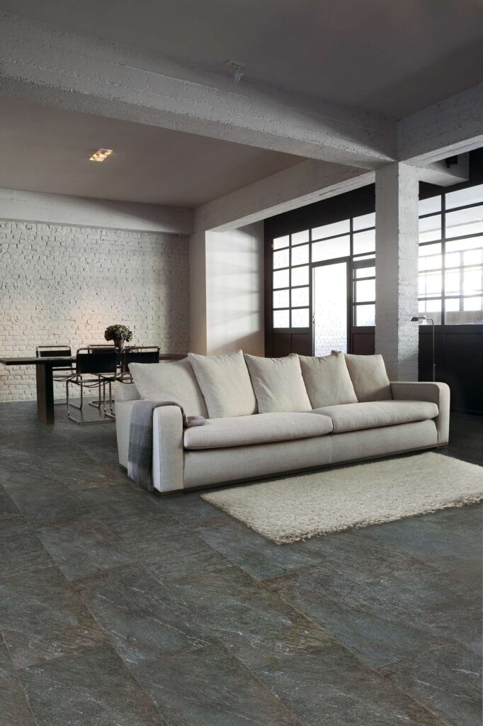 Collezione Discovery in variante Carbon e formato 30x60, utilizzata per il pavimento di un moderno e luminoso ambiente living di design. Le piastrelle in gres porcellanato effetto pietra scura, posate con schema sfalsato, donano carattere e profondità allo spazio, che è arredato con un grande divano color crema, un soffice tappeto chiaro e una parete di mattoni a vista bianchi sullo sfondo, creando un'atmosfera accogliente e dal carattere industriale.