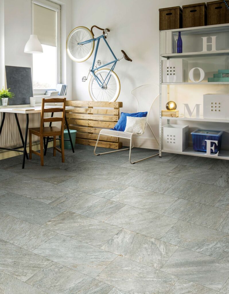 La collezione Discovery Colonial nel formato 30x60 cm riveste con eleganza il pavimento di una luminosa camera da letto con zona studio. Le piastrelle in gres porcellanato effetto pietra naturale, caratterizzate da una texture materica grigia con delicate sfumature calde, sono posate a correre per ampliare visivamente lo spazio. L'ambiente, fresco e giovanile, è valorizzato da una scrivania in legno, una sedia di design in metallo bianco, un'alta scaffalatura a giorno e una bicicletta azzurra appesa alla parete come elemento decorativo.