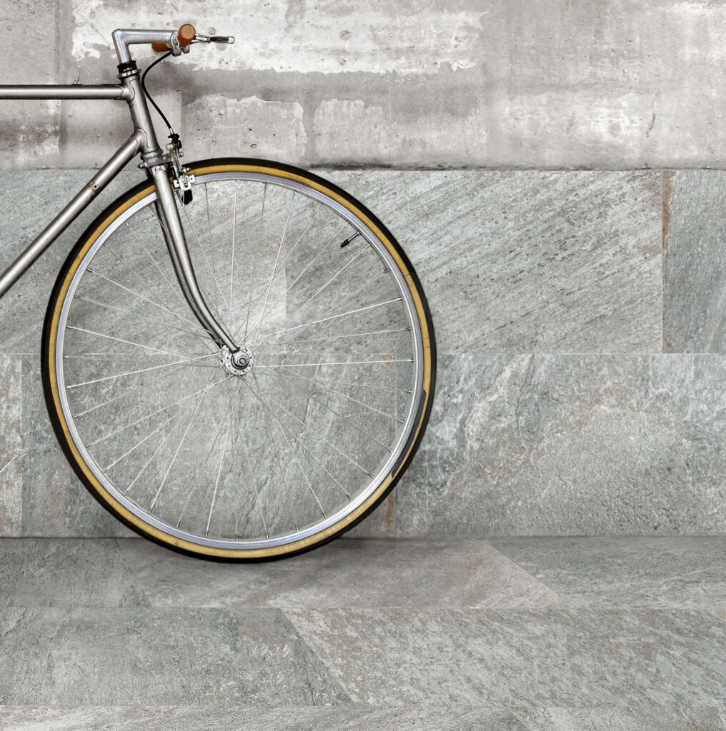 La collezione Discovery Colonial nel formato 30x60 cm valorizza questo spazio d'ispirazione urban-chic con piastrelle in gres porcellanato effetto pietra dalle ricche sfumature grigie e naturali. Nell'immagine, una bicicletta vintage dal telaio metallico è appoggiata a una parete parzialmente rivestita con posa orizzontale, creando una continuità armonica con il pavimento coordinato e mettendo in risalto la texture materica e antiscivolo della superficie.